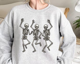 May include: A light gray sweatshirt featuring a graphic of three dancing skeletons. The skeletons are depicted in a detailed, anatomical style, with their arms raised in a celebratory pose. The design is centered on the front of the sweatshirt.