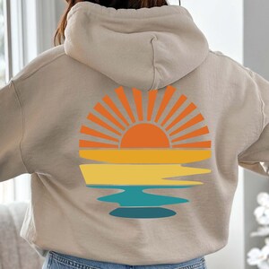 K&ouml;nnte beinhalten: Beiger Kapuzenpullover mit einem Sonnenuntergang-Motiv auf dem R&uuml;cken. Das Design zeigt eine orangefarbene Sonne mit Strahlen, &uuml;ber gelben und blauen horizontalen B&auml;ndern, die eine Strand- oder Ozeanszene andeuten. Der Hoodie hat eine Kapuze mit Kordelzug.
