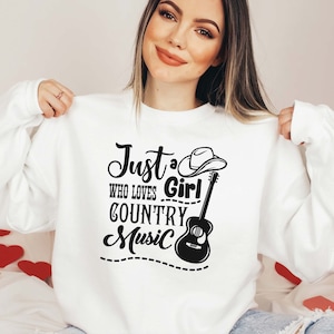 May include: White sweatshirt with black text that reads "Just a Girl Who Loves Country Music". The text is surrounded by a black guitar and a black cowboy hat.