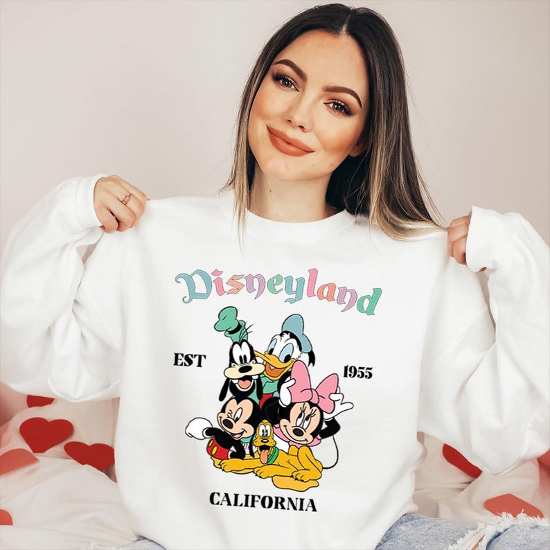 Puede incluir: Sudadera blanca con un gr&aacute;fico de Disneyland con Mickey Mouse, Minnie Mouse, Donald Duck y Goofy. El dise&ntilde;o incluye el texto "Disneyland", "EST 1955" y "CALIFORNIA" en colores pastel. La sudadera es de cuello redondo.