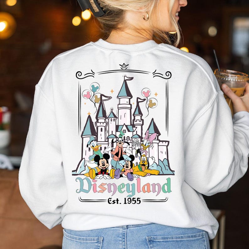 Puede incluir: Sudadera blanca con un gr&aacute;fico de Disneyland. El dise&ntilde;o incluye un castillo, globos y varios personajes de Disney como Mickey Mouse, Donald Duck y Goofy. Tambi&eacute;n est&aacute; presente el texto "Disneyland Est. 1955".