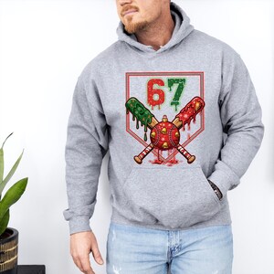 May include: A gray hoodie featuring a baseball-themed design. The graphic includes crossed baseball bats, a baseball ornament, and the number 67 in red and green. The design has a festive, holiday theme.