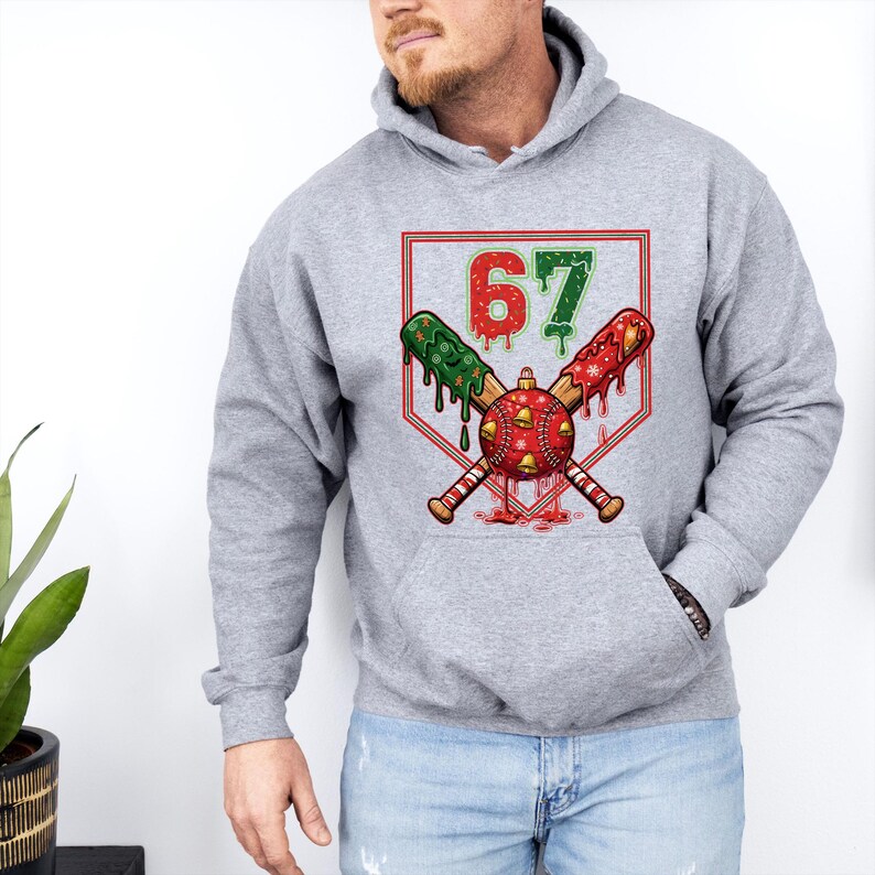 May include: A gray hoodie featuring a baseball-themed design. The graphic includes crossed baseball bats, a baseball ornament, and the number 67 in red and green. The design has a festive, holiday theme.