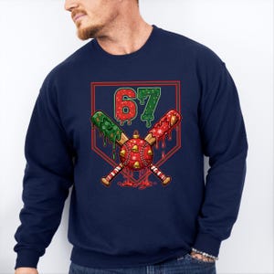 May include: Navy blue sweatshirt featuring a baseball-themed graphic. The design includes crossed baseball bats, a baseball with bells, and the number 67. The bats are decorated with red and green accents, and the number 67 is in red and green.