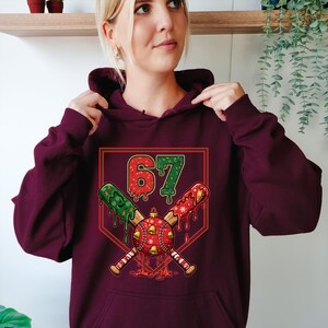 May include: A maroon hoodie with a festive baseball-themed graphic. The graphic features the number "67" in green and red, crossed baseball bats, and a baseball ornament with bells. The design has a Christmas theme.