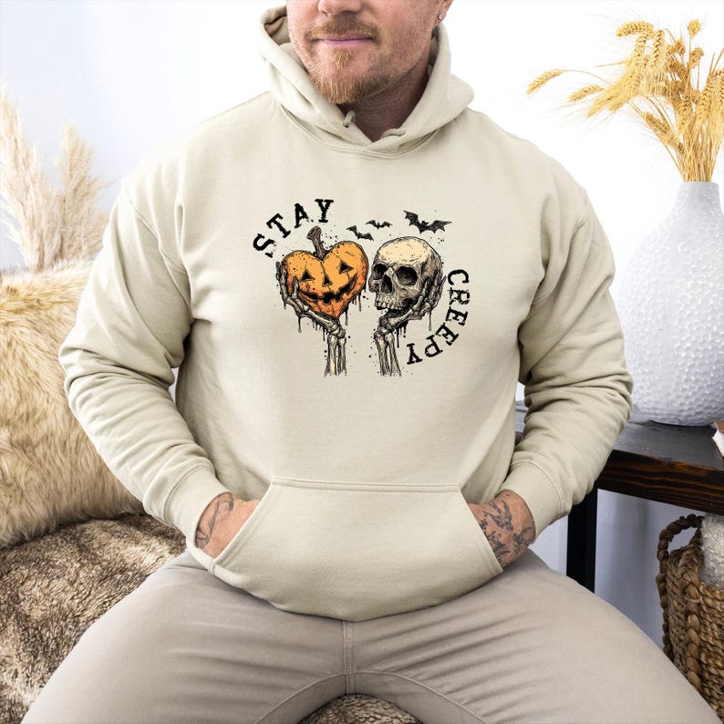 May include: Beige hoodie with a front pocket and a graphic design. The design features a jack-o'-lantern heart and a skull with the words "STAY CREEPY" in a gothic font, along with bat silhouettes. A Halloween-themed apparel item.