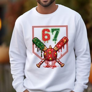 May include: White sweatshirt featuring a baseball-themed Christmas design. The graphic includes crossed baseball bats, a red baseball with bells, and the number "67" in red and green, all within a red and green outlined shape.