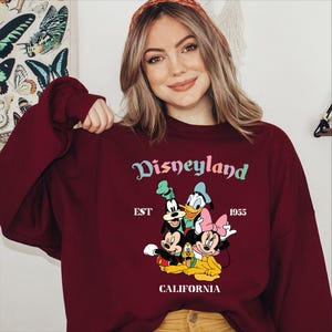 Puede incluir: Sudadera burdeos con la palabra "Disneyland" en una escritura colorida, con Mickey Mouse, Minnie Mouse, Donald Duck y Goofy. Tambi&eacute;n se muestra el texto "EST 1955 CALIFORNIA". Una prenda cl&aacute;sica con tem&aacute;tica de Disney.