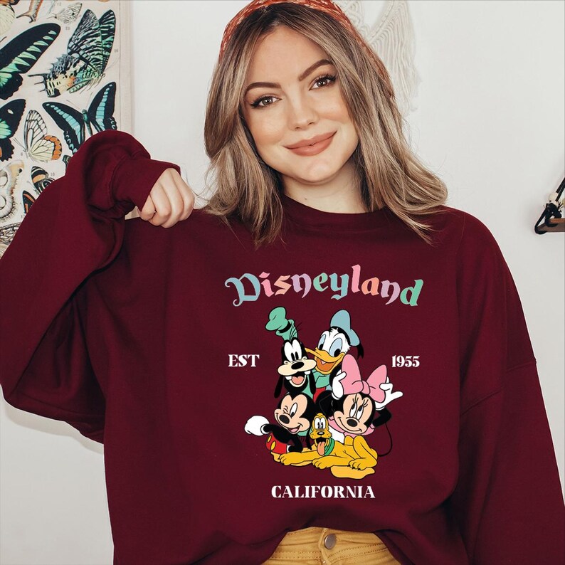 Puede incluir: Sudadera burdeos con la palabra "Disneyland" en una escritura colorida, con Mickey Mouse, Minnie Mouse, Donald Duck y Goofy. Tambi&eacute;n se muestra el texto "EST 1955 CALIFORNIA". Una prenda cl&aacute;sica con tem&aacute;tica de Disney.