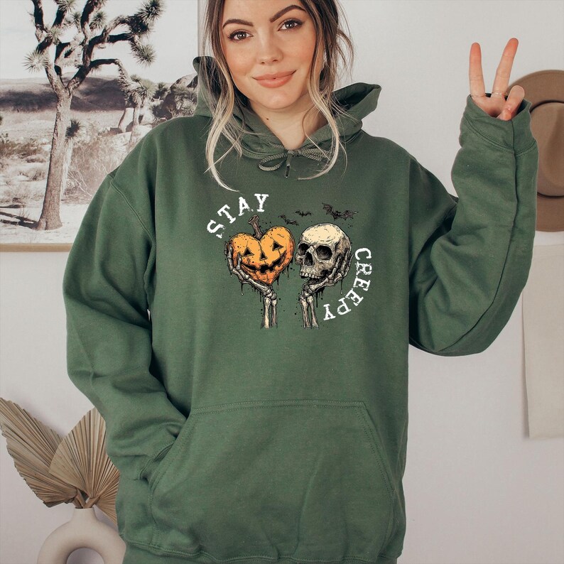 May include: A dark green hoodie with a graphic that says "STAY CREEPY." The graphic features a jack-o'-lantern heart and a skull held by skeleton hands. Two bats are above the skull. The hoodie has a drawstring hood and a front pocket.