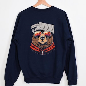 May include: Navy blue sweatshirt featuring a bear wearing sunglasses with a sunset scene reflected in the lenses. The bear also wears a retro-style hat and a red and orange zip-up jacket. The sweatshirt is hanging on a wooden hanger.