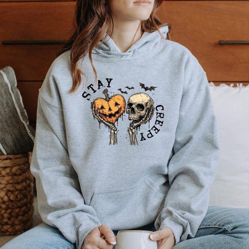 May include: A light gray hoodie with the words "STAY CREEPY" printed in black. The graphic features a jack-o'-lantern heart and a skull held by skeletal hands, with bats above. The hoodie has a hood and a front pocket.