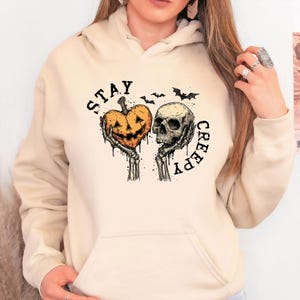 May include: A beige hoodie featuring a Halloween-themed graphic. The design includes a heart-shaped jack-o'-lantern held by skeletal hands, a skull, and the words "STAY CREEPY" in a circular arrangement, with bats above.