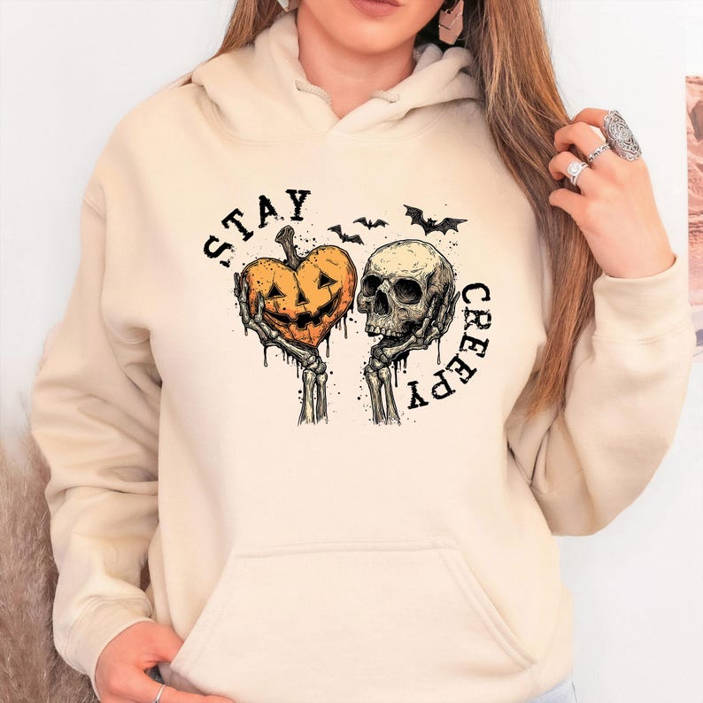 May include: A beige hoodie featuring a Halloween-themed graphic. The design includes a heart-shaped jack-o'-lantern held by skeletal hands, a skull, and the words "STAY CREEPY" in a circular arrangement, with bats above.