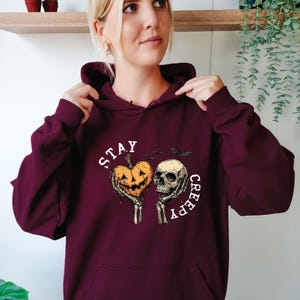May include: Burgundy hoodie with a Halloween-themed graphic. The design features a jack-o'-lantern heart and a skull held by skeletal hands, with the words "STAY CREEPY" in a circular layout. Two bats are also included in the design.