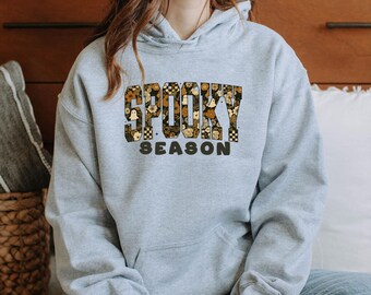 May include: A light gray hoodie with the words "SPOOKY SEASON" in a decorative, patterned font. The text features a mix of brown, black, and white elements, including floral and checkerboard designs. The hoodie has a drawstring hood and a kangaroo pocket.