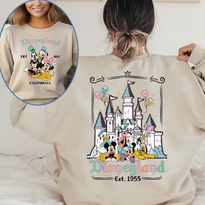 Puede incluir: Sudadera beige con gr&aacute;ficos de Disneyland en la parte delantera y trasera. El gr&aacute;fico frontal incluye a Mickey, Donald y Minnie Mouse, con el texto "Disneyland EST 1955 CALIFORNIA". El gr&aacute;fico trasero muestra un castillo y personajes de Disney.