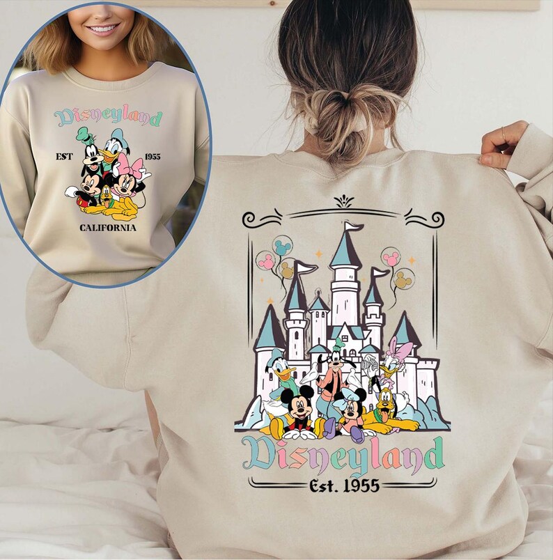 Puede incluir: Sudadera beige con gr&aacute;ficos de Disneyland en la parte delantera y trasera. El gr&aacute;fico frontal incluye a Mickey, Donald y Minnie Mouse, con el texto "Disneyland EST 1955 CALIFORNIA". El gr&aacute;fico trasero muestra un castillo y personajes de Disney.