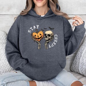 May include: Dark gray hoodie with a Halloween-themed graphic. The design features a jack-o'-lantern heart and a skull held by skeleton hands, with the words "STAY CREEPY" in a circular arrangement. Two bats are also included.
