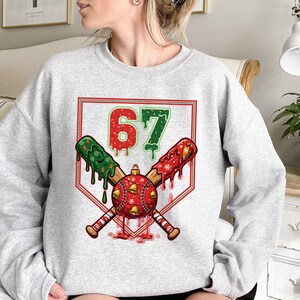 May include: A light gray sweatshirt featuring a baseball-themed Christmas design. The graphic includes crossed baseball bats, a baseball ornament, and the number 67 in red and green. The design has a festive, holiday theme.