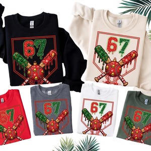 May include: A collection of sweatshirts in various colors, including black, red, white, and gray. Each sweatshirt features a graphic of a baseball with the number 67, crossed bats, and festive dripping accents. The design is set within a diamond shape.