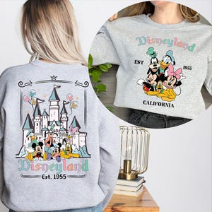 Puede incluir: Sudadera gris claro con un dise&ntilde;o de Disneyland. La parte trasera muestra un castillo y personajes, mientras que la parte delantera muestra a Mickey, Donald y Minnie Mouse. El texto "Disneyland Est. 1955" est&aacute; en colores pastel.