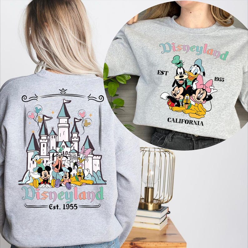 Puede incluir: Sudadera gris claro con un dise&ntilde;o de Disneyland. La parte trasera muestra un castillo y personajes, mientras que la parte delantera muestra a Mickey, Donald y Minnie Mouse. El texto "Disneyland Est. 1955" est&aacute; en colores pastel.