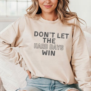 May include: A beige sweatshirt with the text "DON'T LET THE HARD DAYS WIN" in dark gray letters. The long-sleeved top has a crew neck and a relaxed fit. The sweatshirt is made of a soft material.