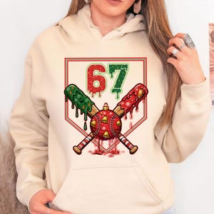 May include: Cream-colored hoodie featuring a baseball-themed graphic. The design includes crossed baseball bats, a baseball with bells, and the number 67 in red and green. The bats and number have a dripping effect, with red and green accents.