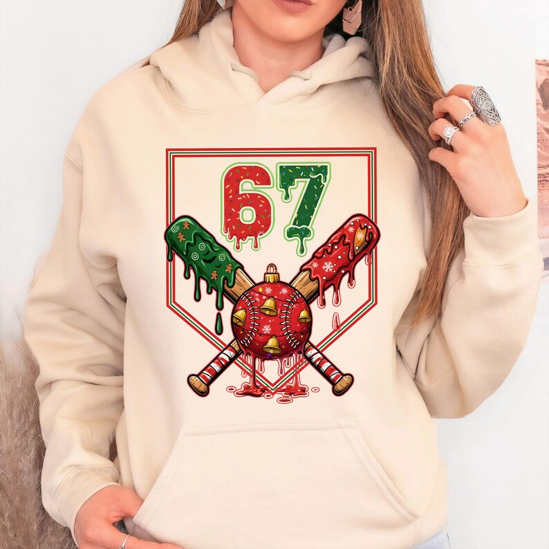 May include: Cream-colored hoodie featuring a baseball-themed graphic. The design includes crossed baseball bats, a baseball with bells, and the number 67 in red and green. The bats and number have a dripping effect, with red and green accents.