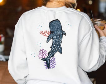 May include: White sweatshirt with a blue whale shark illustration on the back. The shark is surrounded by coral and seaweed in shades of pink, purple, and green.