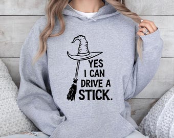 May include: A light gray hoodie with a black graphic of a witch's hat and broomstick. The text on the hoodie reads "YES I CAN DRIVE A STICK."