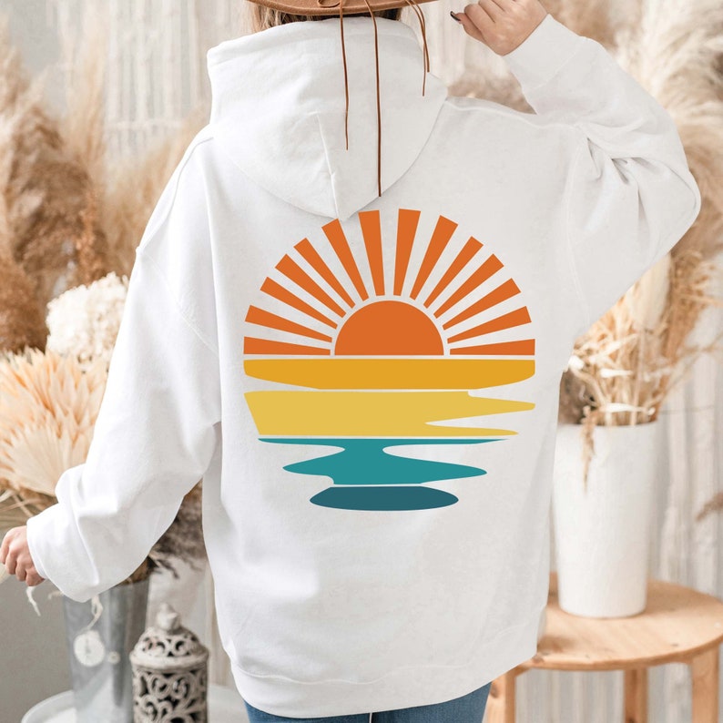 K&ouml;nnte beinhalten: Wei&szlig;er Kapuzenpullover mit einem Sonnenuntergangsdesign auf dem R&uuml;cken. Die Grafik zeigt eine Sonne mit orangefarbenen und gelben Strahlen &uuml;ber gelben, orangefarbenen und t&uuml;rkisfarbenen horizontalen Streifen, die an ein Strand- oder Ozeanthema erinnern.