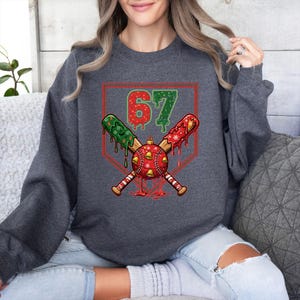 May include: Dark gray sweatshirt featuring a baseball-themed Christmas design. The graphic includes crossed baseball bats, a red and green baseball ornament, and the number 67 in green. The design is set within a red baseball home plate.