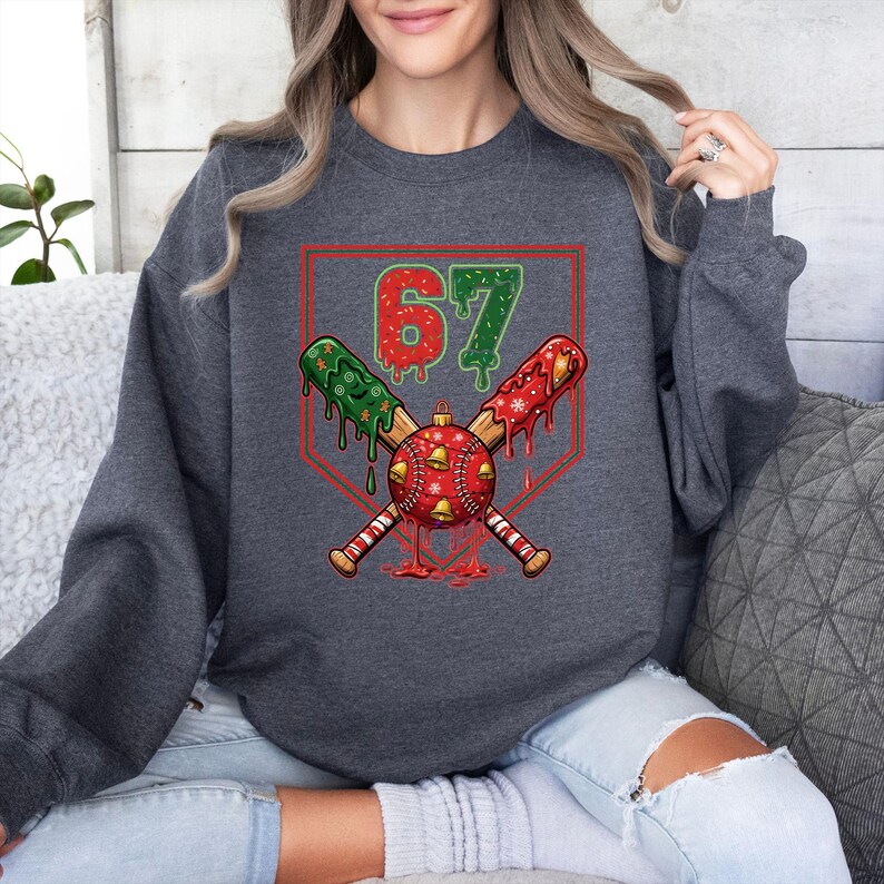 May include: Dark gray sweatshirt featuring a baseball-themed Christmas design. The graphic includes crossed baseball bats, a red and green baseball ornament, and the number 67 in green. The design is set within a red baseball home plate.