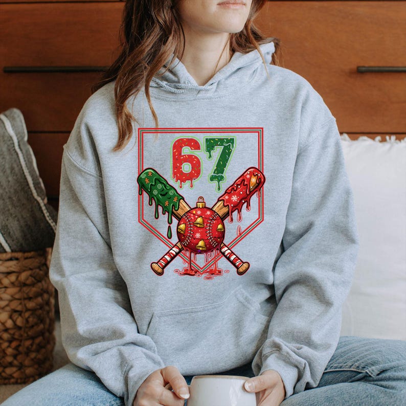 May include: A light gray hoodie featuring a baseball-themed graphic. The graphic includes the number 67, crossed baseball bats, and a baseball with Christmas decorations. The bats are green and red with dripping accents.