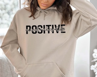 May include: A beige hoodie with a front pocket and drawstring hood. The word "POSITIVE" is printed in bold black letters across the chest, with "mind vibes life" in a script font underneath.