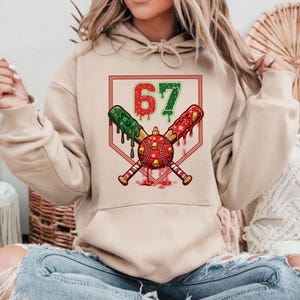May include: Beige hoodie featuring a baseball-themed Christmas graphic. The design includes crossed baseball bats, a red and green baseball, and the number 67. The bats have green and red accents, and the baseball is decorated with bells.
