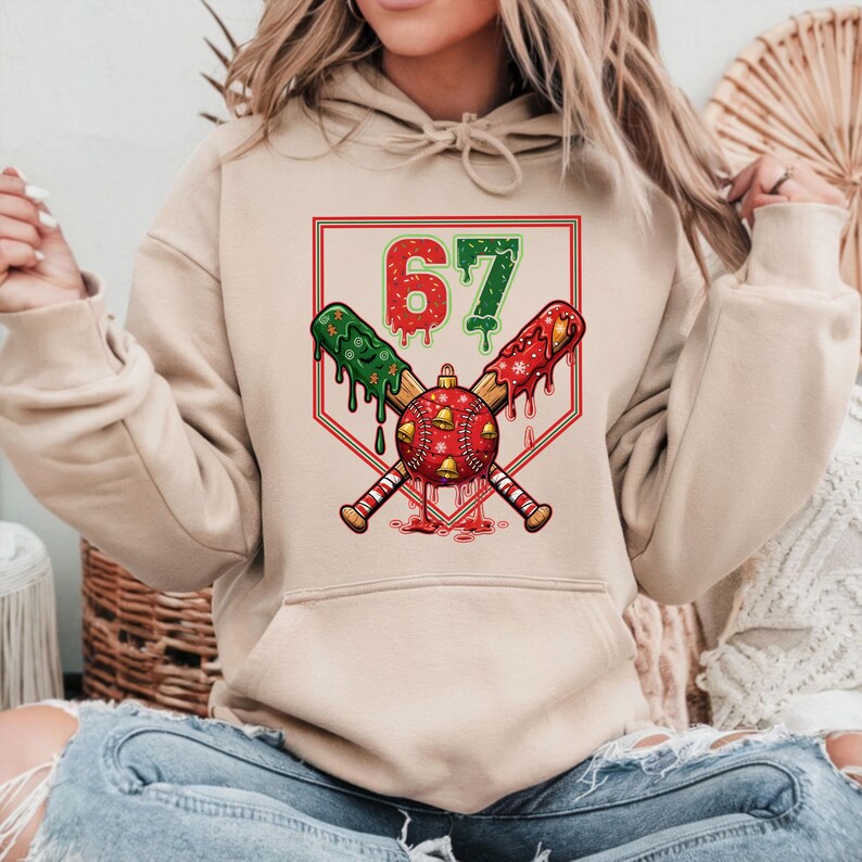 May include: Beige hoodie featuring a baseball-themed Christmas graphic. The design includes crossed baseball bats, a red and green baseball, and the number 67. The bats have green and red accents, and the baseball is decorated with bells.