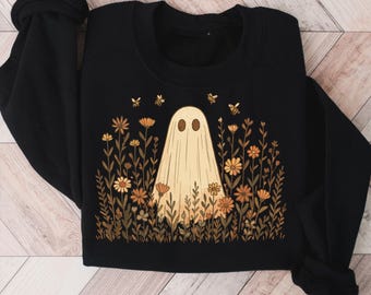 May include: Black sweatshirt featuring a beige ghost design surrounded by flowers and bees. The ghost has oval eyes and is standing in a field of flowers in shades of orange, yellow, and brown. The bees are yellow and black.