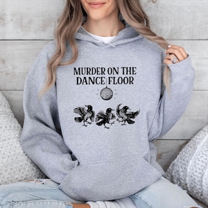 May include: A light gray hoodie with the text "MURDER ON THE DANCE FLOOR" in black. Below the text is a disco ball graphic, and three black raven illustrations. The hoodie has a drawstring hood and a front pocket.