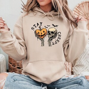May include: Beige hoodie with a Halloween-themed graphic. The design features a jack-o'-lantern heart held by skeletal hands next to a skull, with the words "STAY CREEPY" in a distressed font. Includes bats.