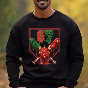 May include: Black sweatshirt featuring a festive baseball-themed graphic. The design includes crossed baseball bats, a red and gold ornament with bells, and the number "67" in red and green. The bats and ornament have a dripping effect.
