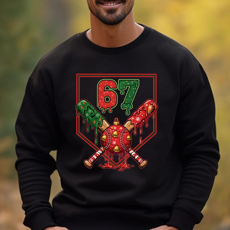 May include: Black sweatshirt featuring a festive baseball-themed graphic. The design includes crossed baseball bats, a red and gold ornament with bells, and the number "67" in red and green. The bats and ornament have a dripping effect.