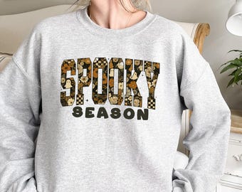May include: A light gray sweatshirt with the words "SPOOKY SEASON" in a floral and checkered design. The word "SPOOKY" is in a bold, block letter style, with a ghost graphic. The word "SEASON" is in a simple, sans-serif font.