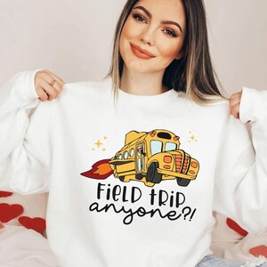 May include: White sweatshirt with a yellow school bus with flames coming out the back. The bus is flying through the air with stars around it. The text on the sweatshirt says "Field trip anyone?"