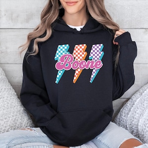 May include: Black hoodie featuring a graphic of three lightning bolts with checkered patterns in teal, orange, and purple. The word "Boone" is written in a pink script font across the lightning bolts.