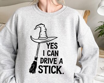May include: A light gray sweatshirt featuring a black witch hat and broom graphic. The text "YES I CAN DRIVE A STICK" is printed below the graphic in a bold, black font. The sweatshirt is a casual, comfortable style.