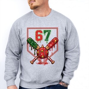 May include: A light gray sweatshirt featuring a baseball-themed Christmas design. The graphic includes crossed baseball bats, a red and gold ornament, and the number "67" in red and green. The bats have green and red accents.