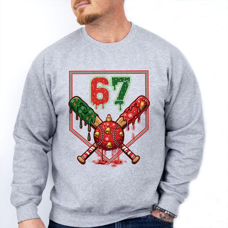 May include: A light gray sweatshirt featuring a baseball-themed Christmas design. The graphic includes crossed baseball bats, a red and gold ornament, and the number "67" in red and green. The bats have green and red accents.